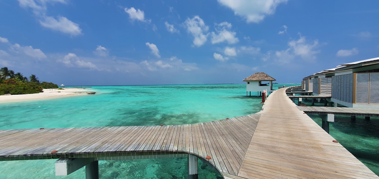 Wooden boardwalk and overwater bungalows at Cinnamon Hakuraa Huraa Maldives with turquoise lagoon, sandy beach, and blue sky.