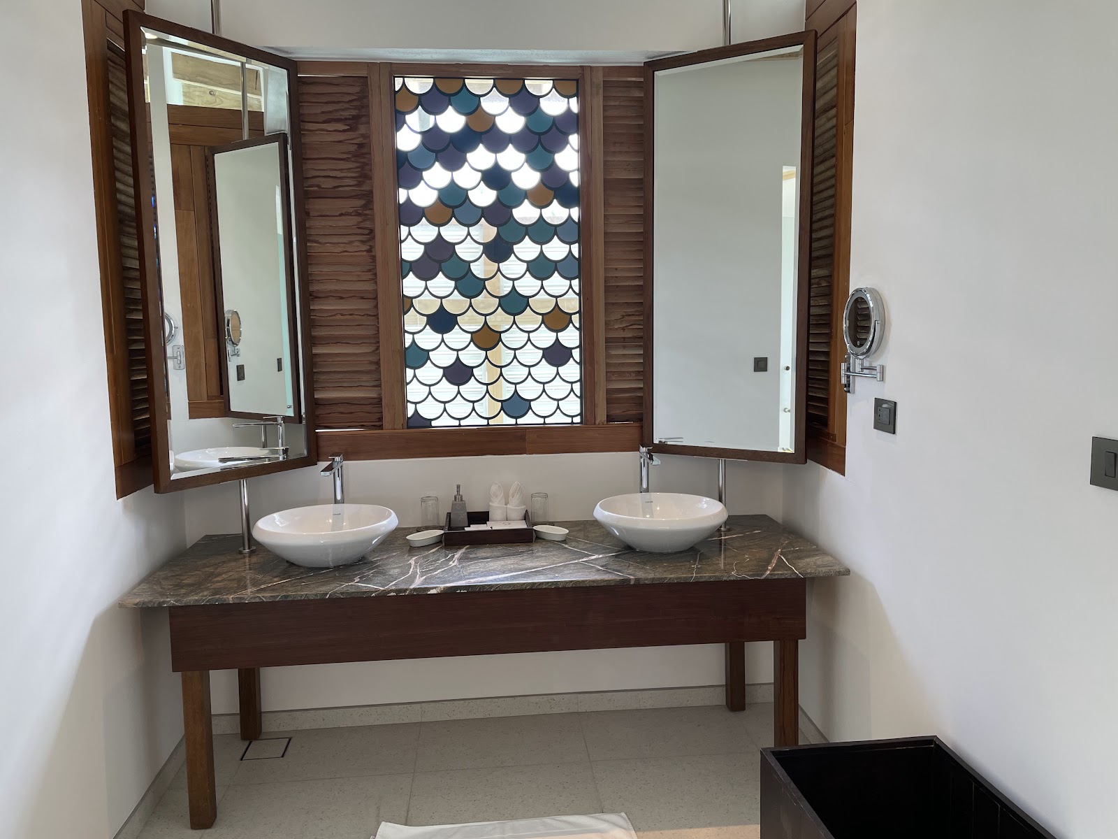 Bathroom with double vessel sinks on dark marble countertop, wooden mirrored cabinets, and decorative scalloped tile window at Cinnamon Hakuraa Huraa Maldives.
