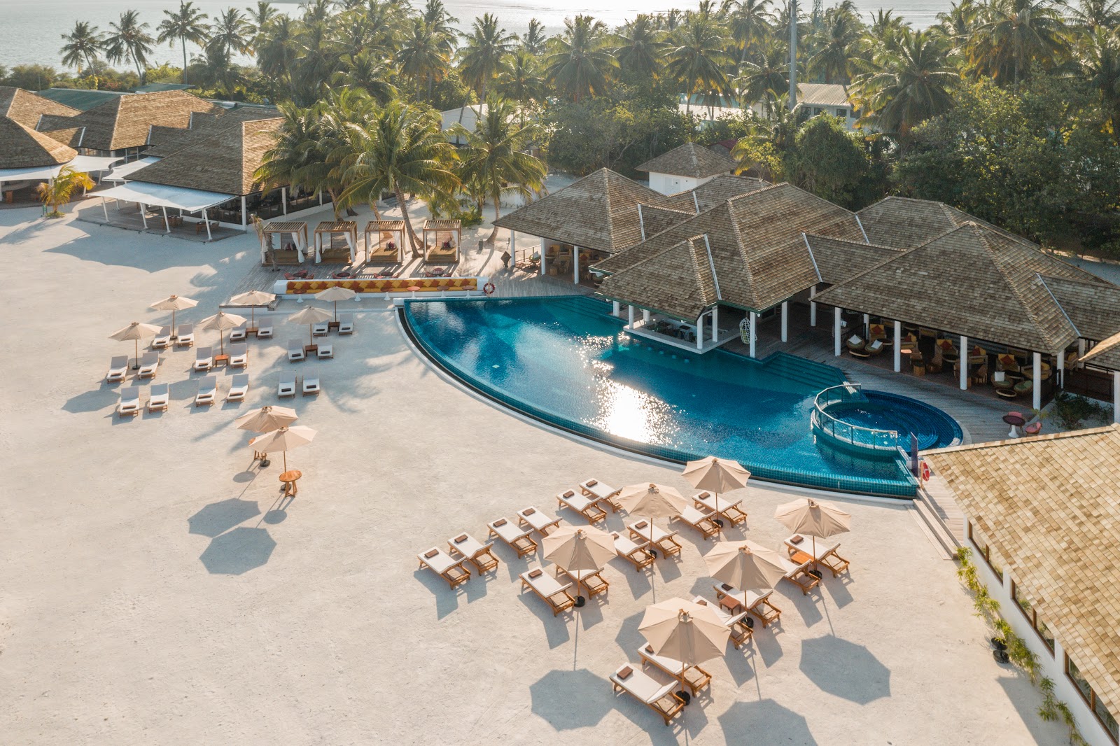 A beachfront bungalow with blue and white exterior and sail-shaped canopy at dusk, with lounge chairs and lanterns on the sand facing calm ocean waters.