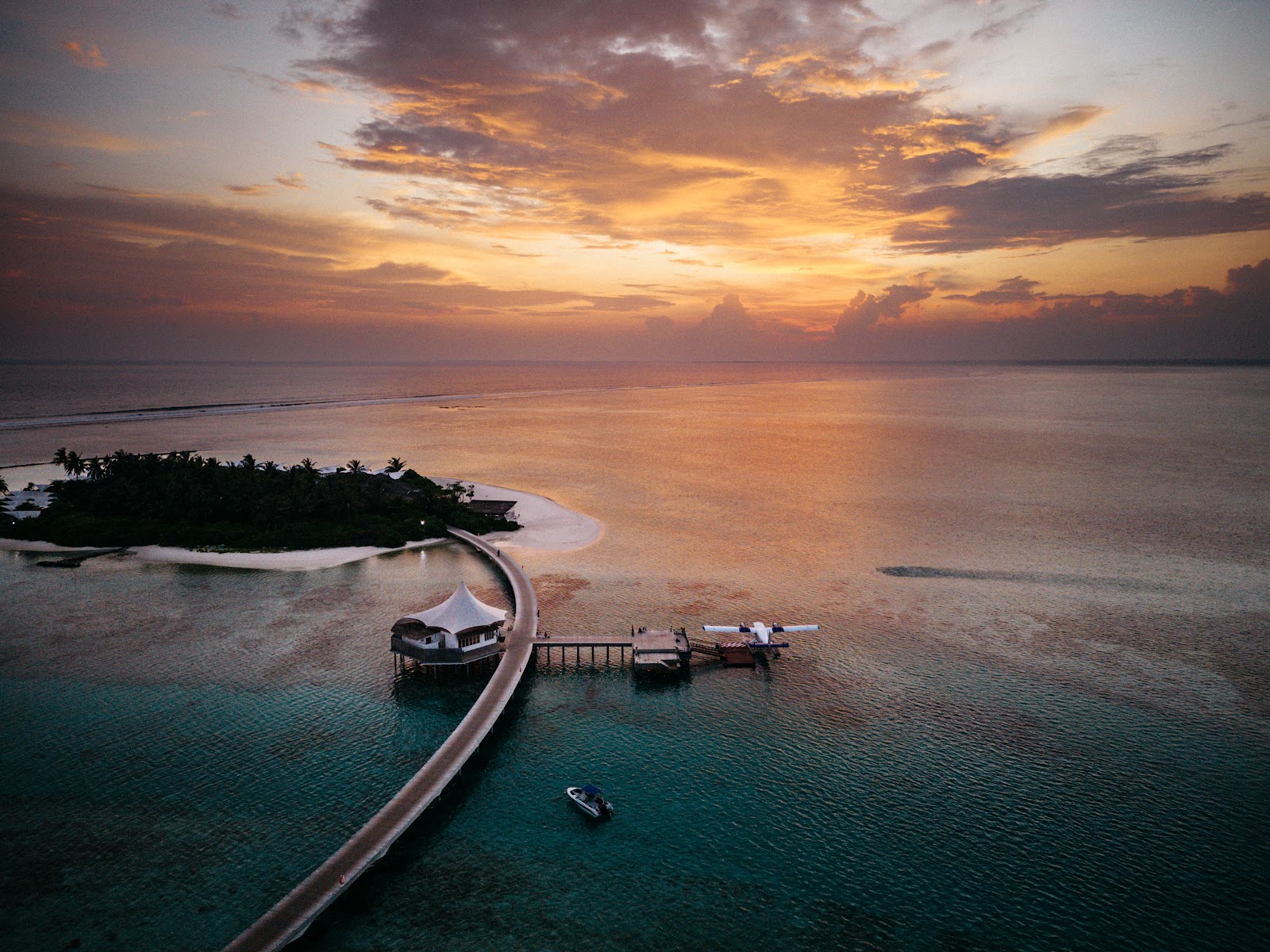 Aerial view of a curved lagoon-style pool with lounge chairs and umbrellas on white sand, flanked by thatched-roof buildings and coconut palms at Cinnamon Hakuraa Huraa Maldives.