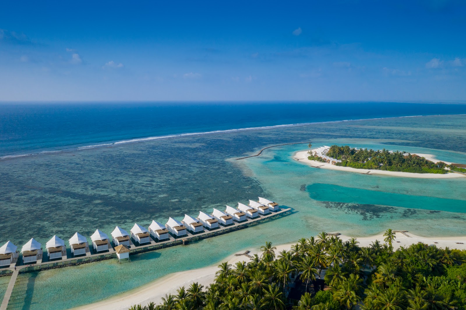 Curved causeway connecting a palm-fringed island to overwater structures at sunset, with turquoise lagoon and golden sky above.