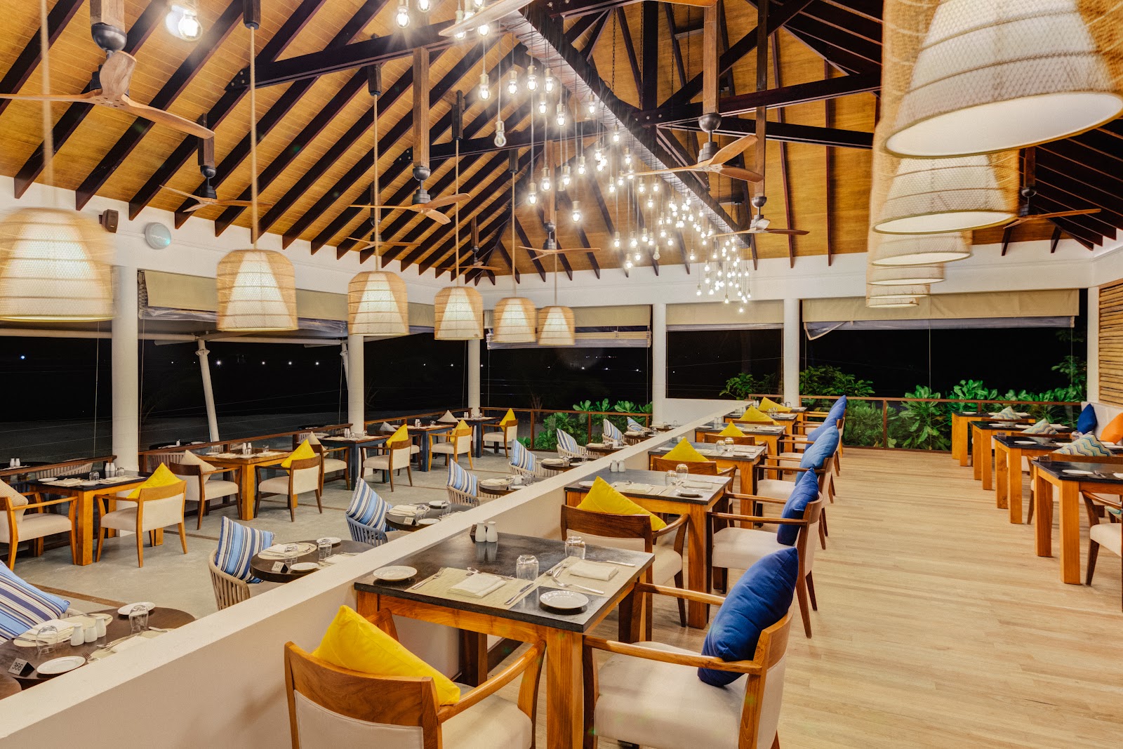 Open-air dining pavilion with wooden beams, pendant lights, and tables set with yellow cushions overlooking the ocean at night.