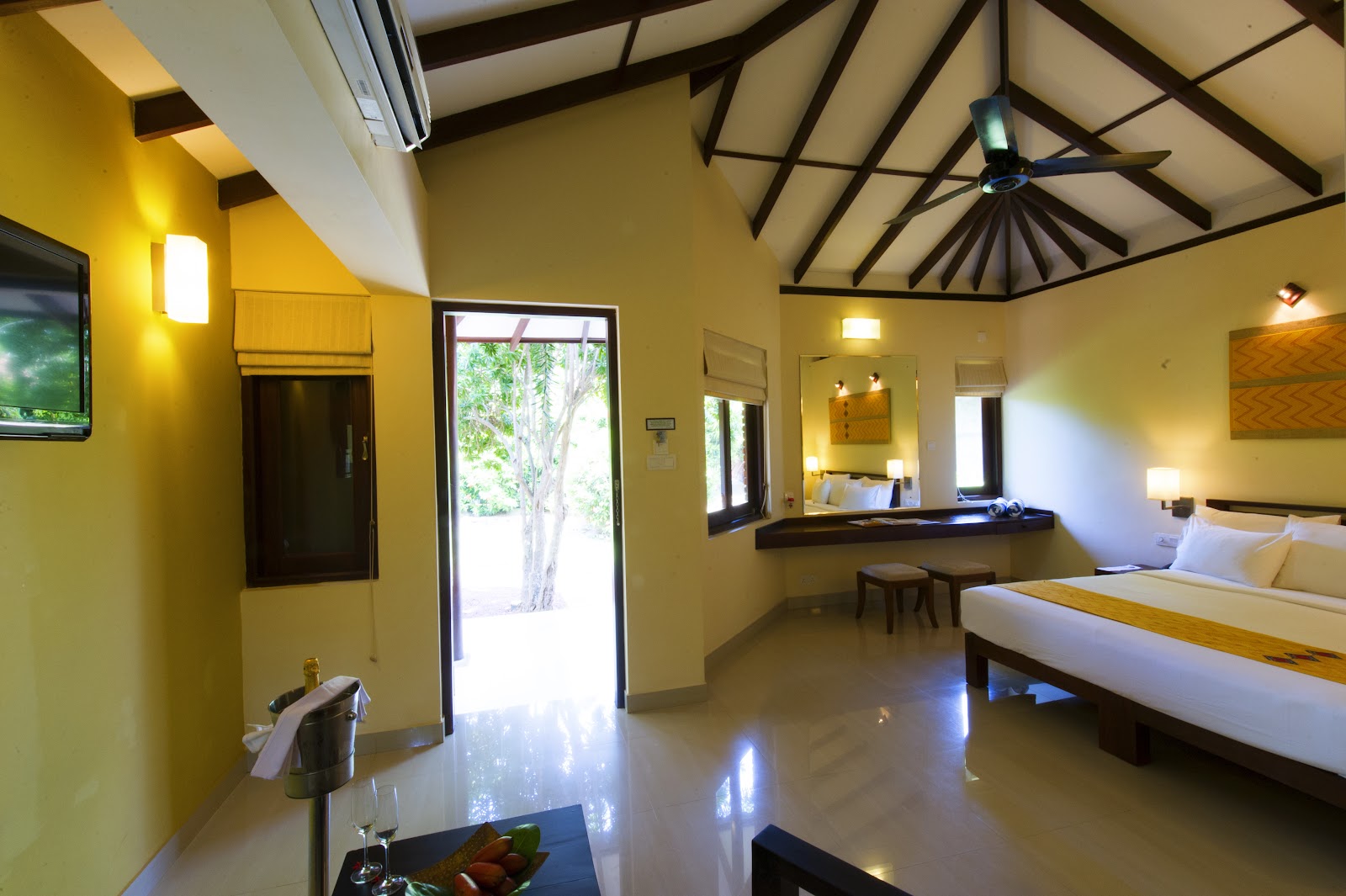 Bright yellow resort room with high vaulted ceiling, ceiling fan, queen bed with gold runner, dark wood furnishings, and glass doors opening to tropical grounds.
