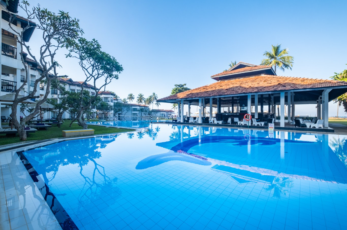 Large blue swimming pool with curved lanes at Club Hotel Dolphin, flanked by white buildings, a covered pavilion, and palm trees under clear sky.