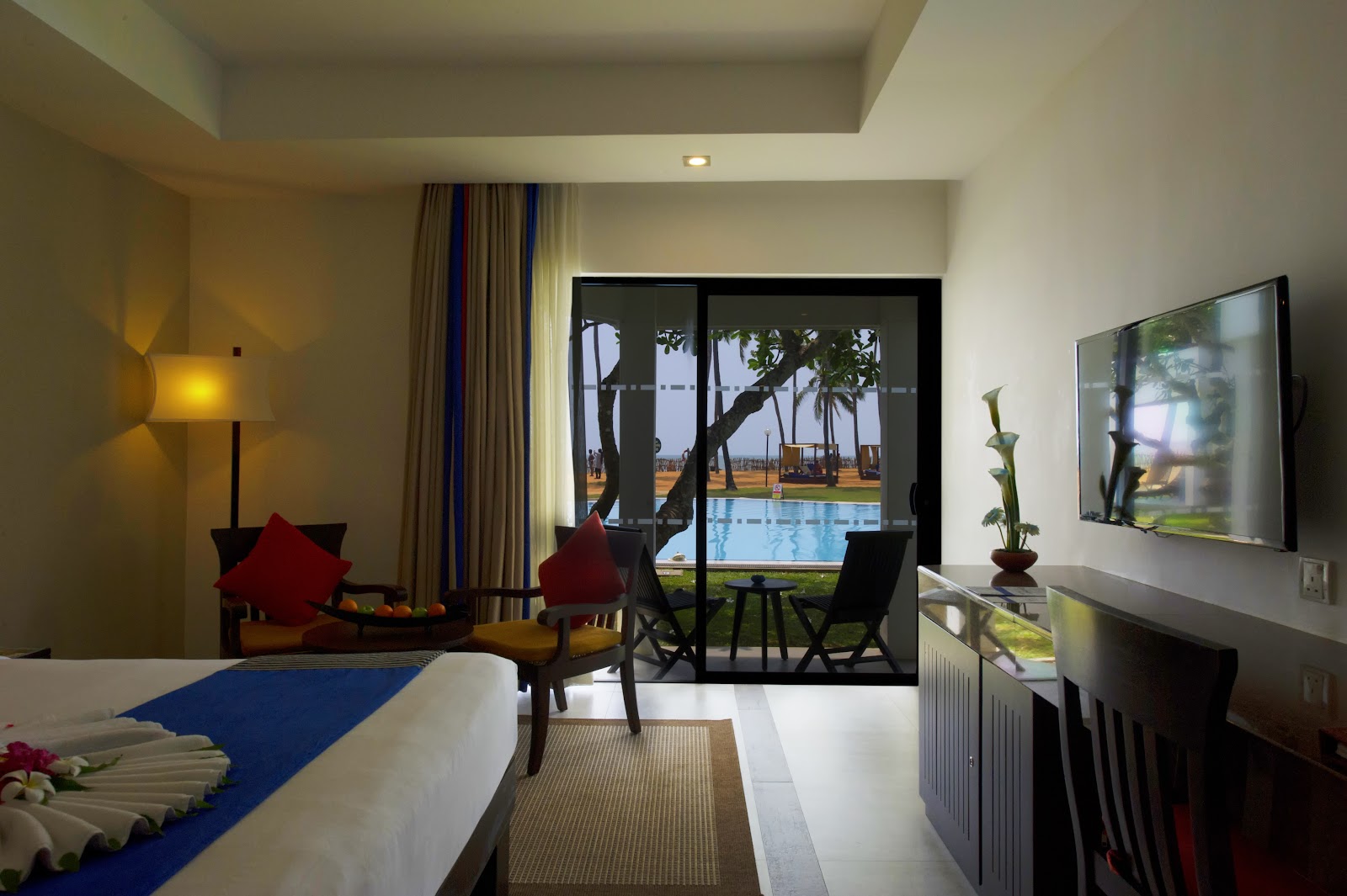 Resort room with bed, seating area with red cushions, and glass doors opening to pool and palm trees beyond.