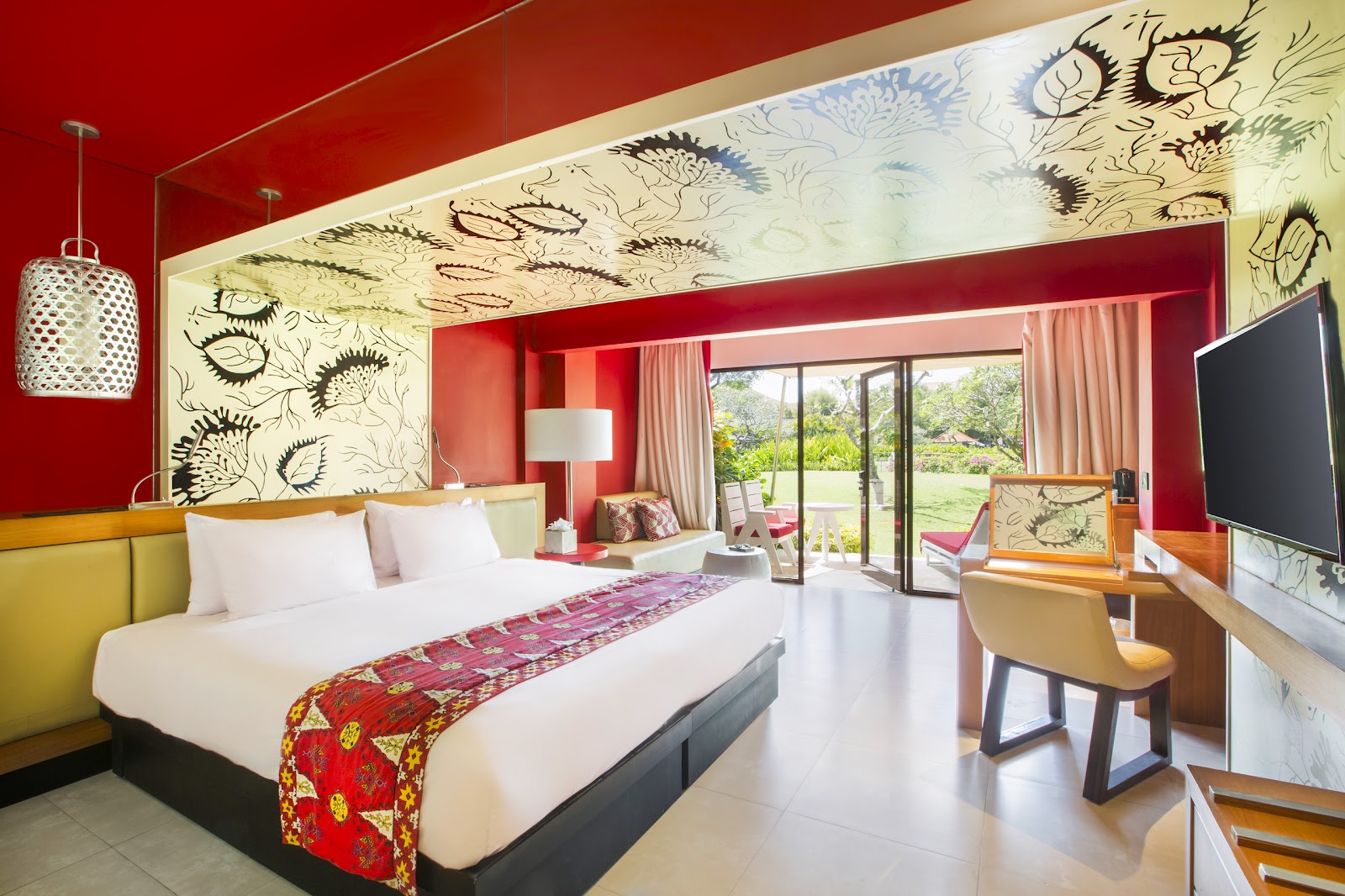 Bright resort bedroom with red walls, white bed, patterned ceiling, and glass doors opening to gardens and pool area.