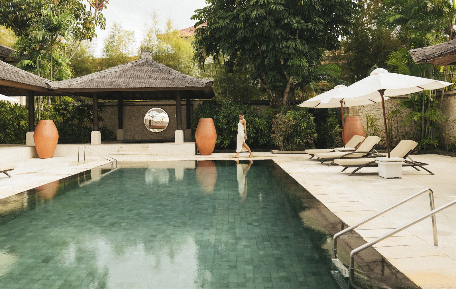 Pool area surrounded by loungers and white umbrellas, with a thatched pavilion and large terra-cotta urns amid lush tropical vegetation.