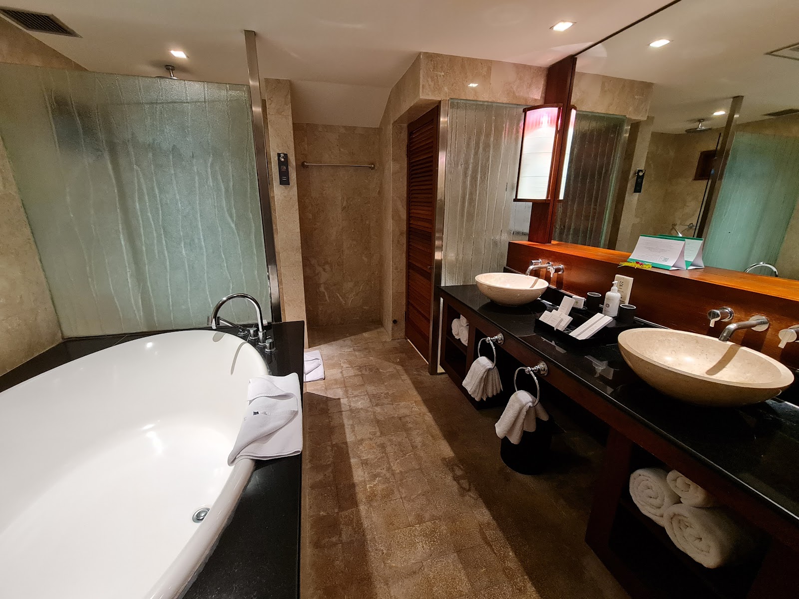 Spacious bathroom with freestanding soaking tub on left, dual vessel sinks on dark wood vanity, walk-in shower with glass enclosure, and rolled towels on shelves.