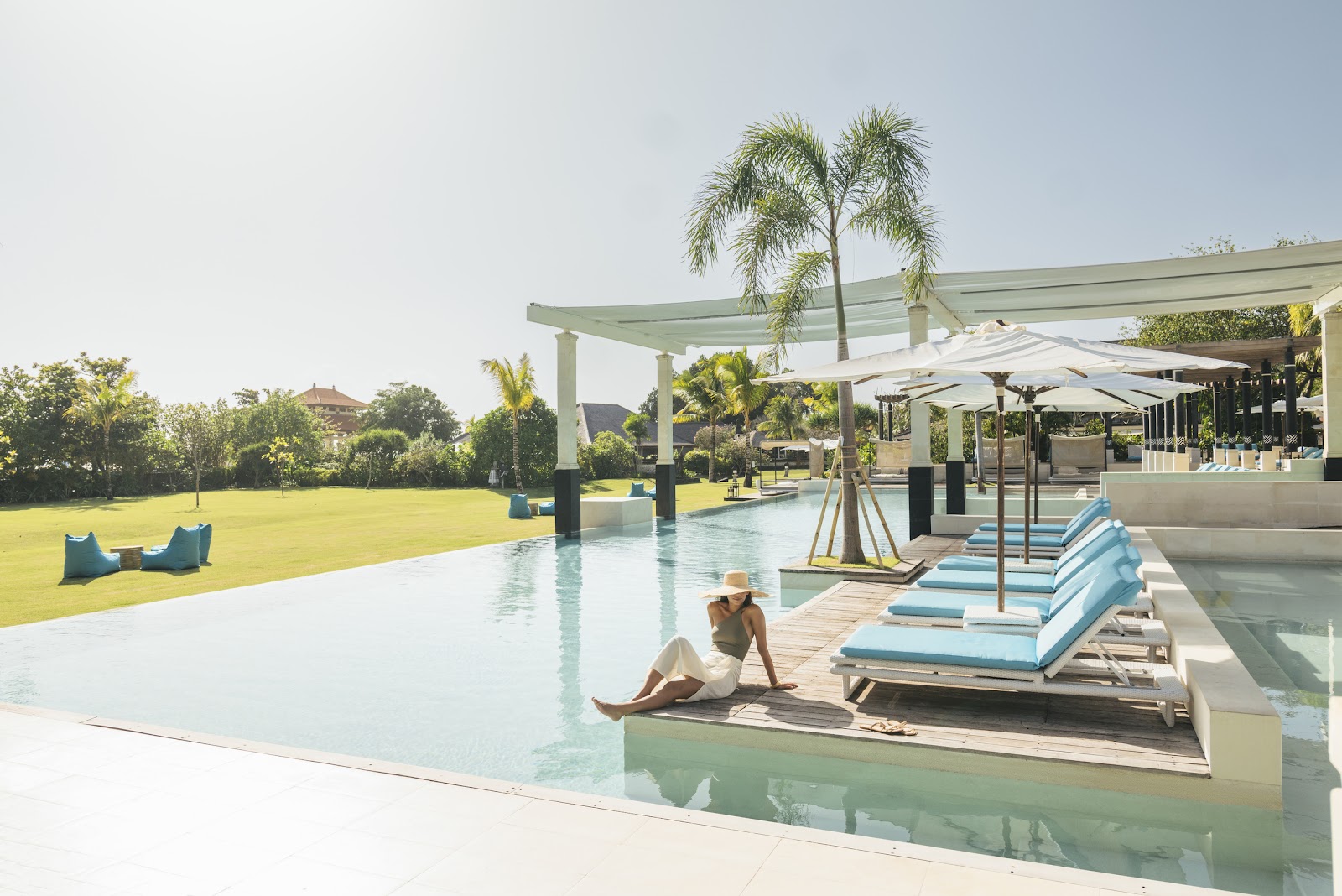 Resort guest relaxing on a blue lounger beside a large pool with palm trees, white umbrellas, and manicured lawn at Club Med Bali.