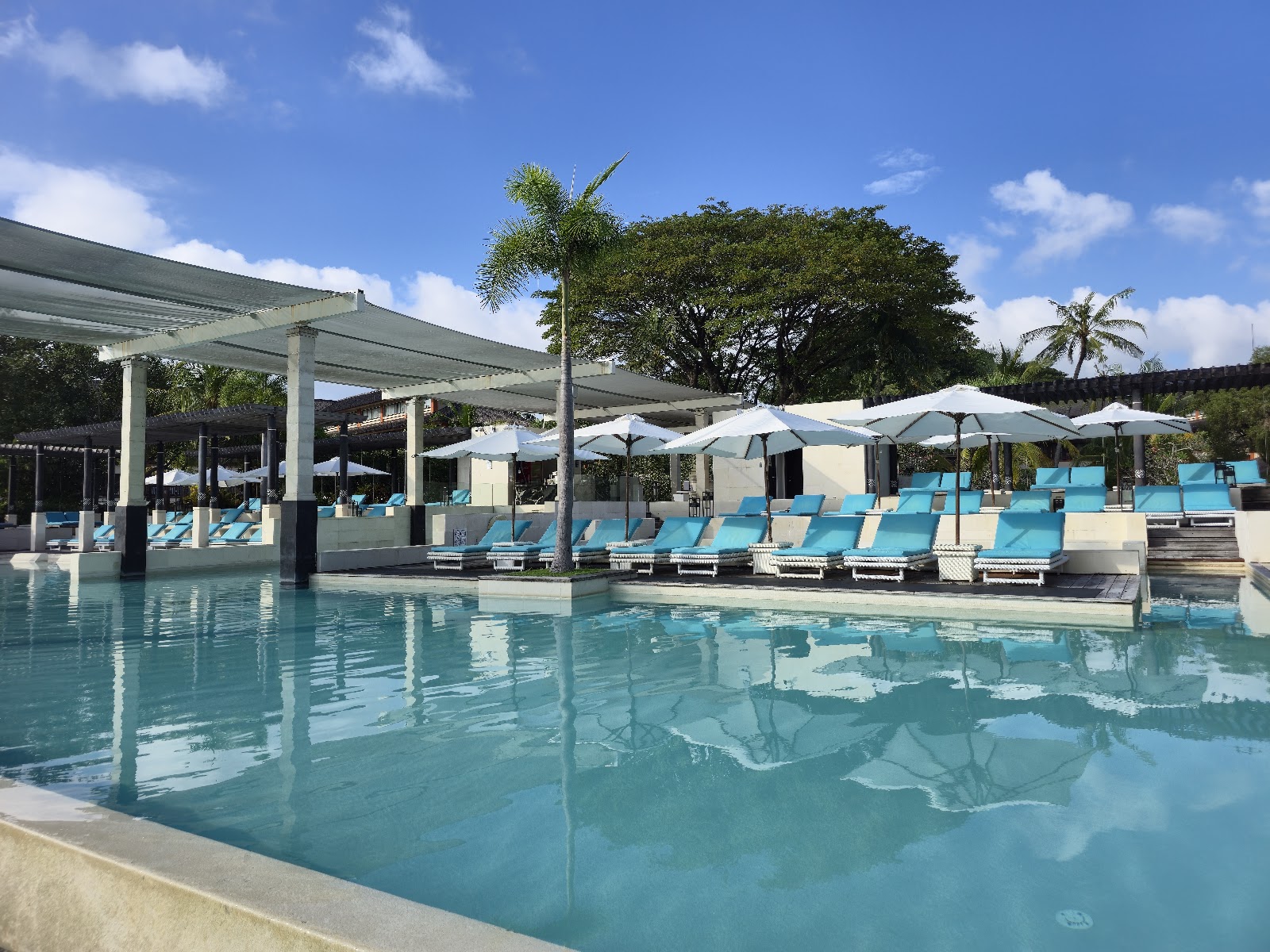 Resort pool area with turquoise water, white pergolas, blue lounge chairs, umbrellas, and tropical trees under clear sky.
