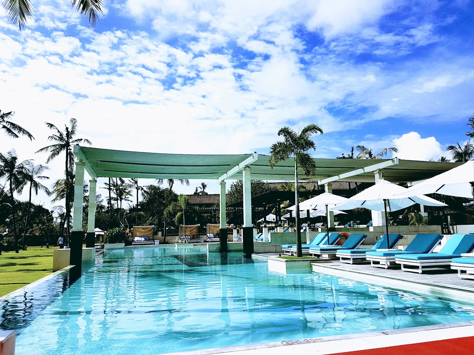 Large resort pool with green pergolas, blue loungers, white umbrellas, and palm trees under a partly cloudy sky.