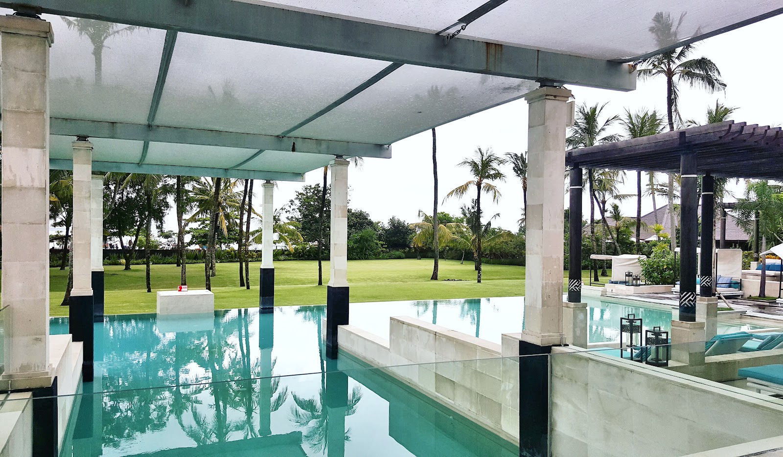 Turquoise pool under a glass-roofed pavilion with palm trees and manicured lawns visible beyond the covered structure.
