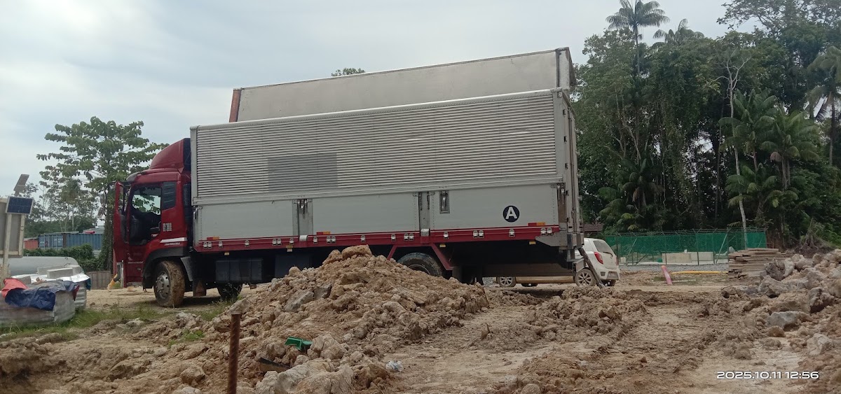 Red and white delivery truck parked on sandy construction site surrounded by palm trees and earthen materials.