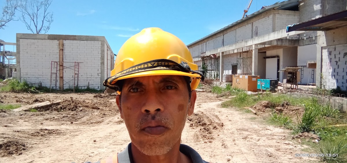 Construction worker wearing a yellow hard hat on an active building site with concrete structures and bare ground.