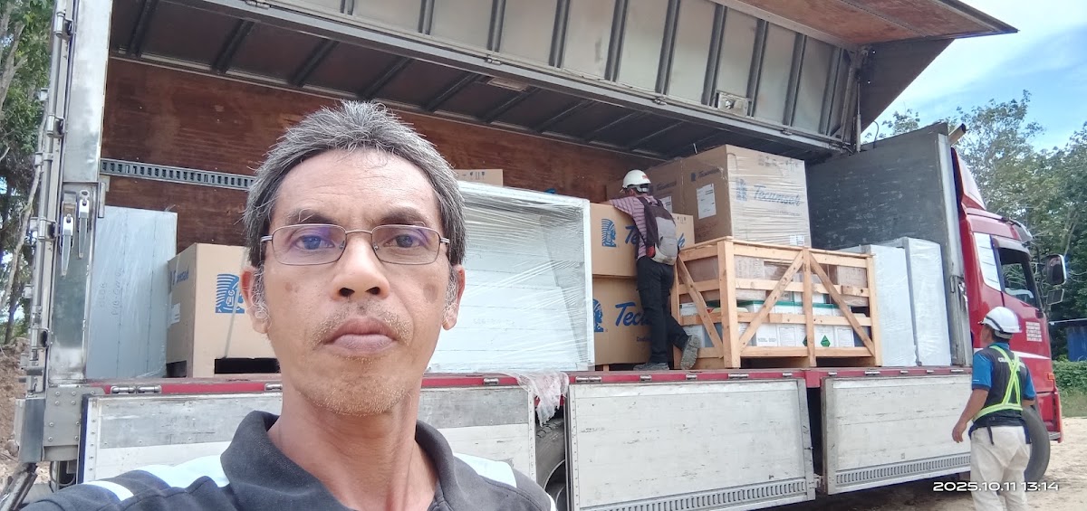 Man wearing glasses standing in front of an open delivery truck loaded with cardboard boxes and wooden crates.