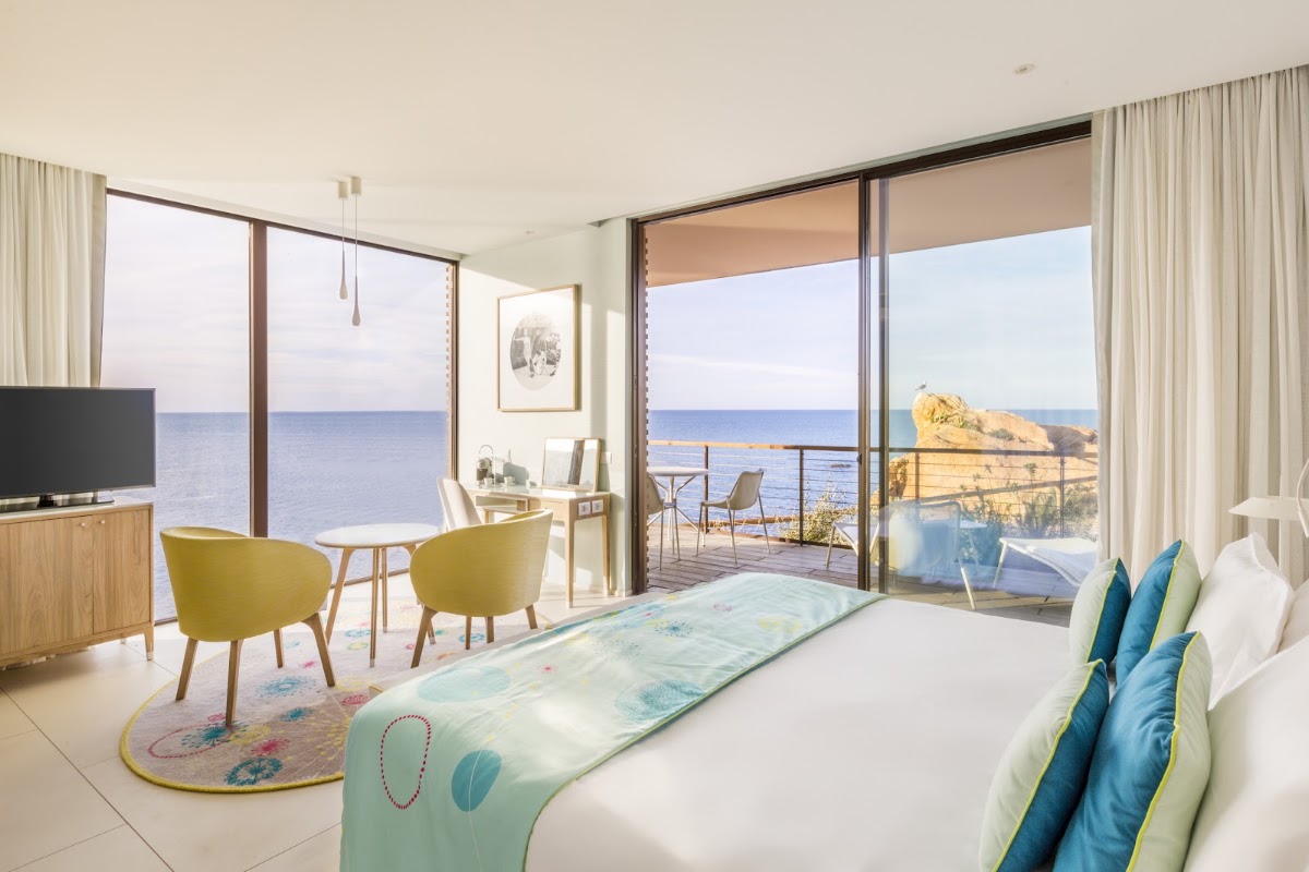 Resort bedroom with sea view, bed with blue and green pillows, yellow chairs, and private balcony overlooking Mediterranean coastline and rocky outcrop.