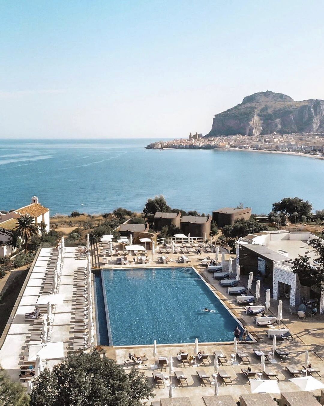 Resort pool with lounge chairs and umbrellas overlooks turquoise Mediterranean waters and the coastal town of Cefalù backed by dramatic cliffs.