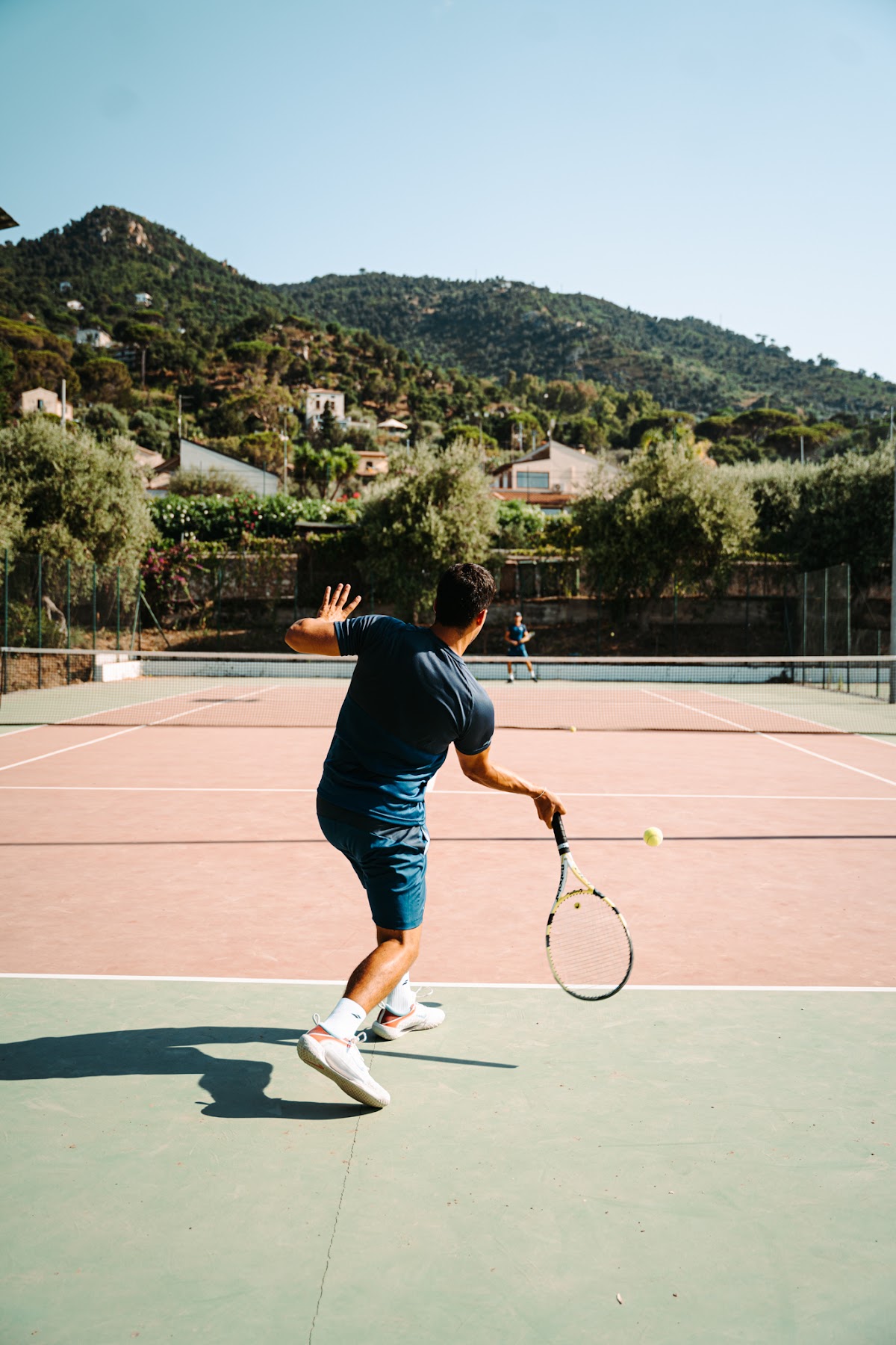 Man playing tennis on clay court at Club Med Cefalù with forested hills and Mediterranean villas in the background.