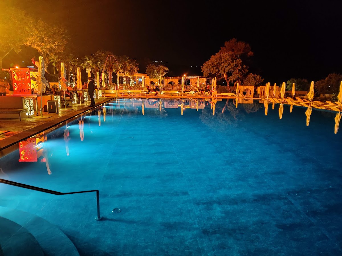 Illuminated pool at night with turquoise water, white umbrellas lining both sides, golden-lit buildings and palm trees in the background.