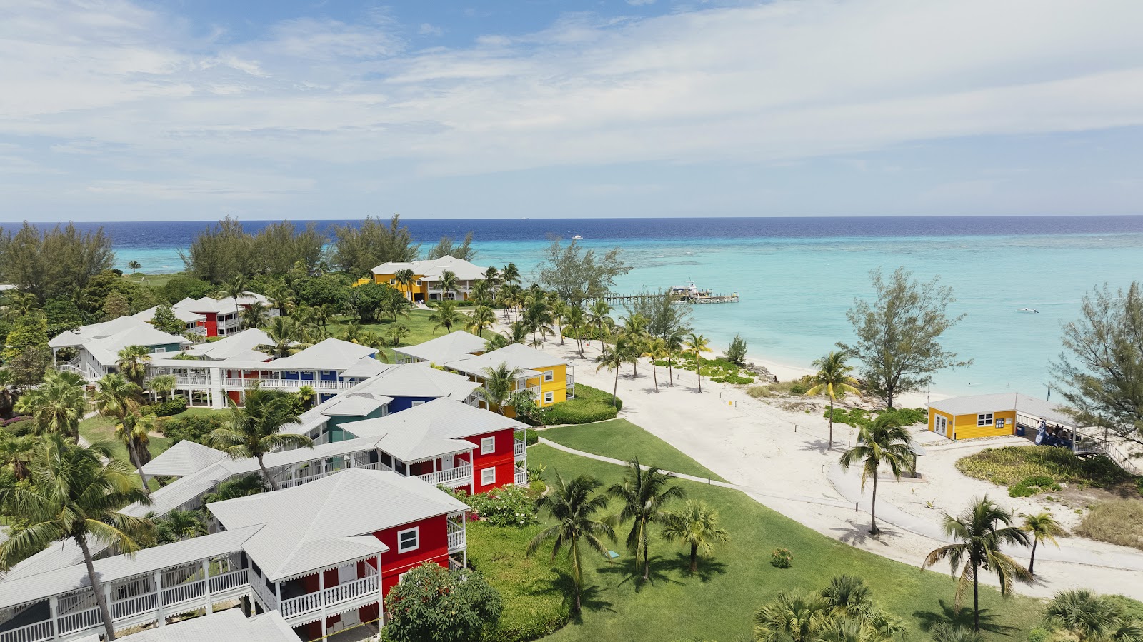 Colorful beachfront cottages at Club Med Columbus Isle overlook turquoise waters, white sand, and a wooden pier on a tropical island.