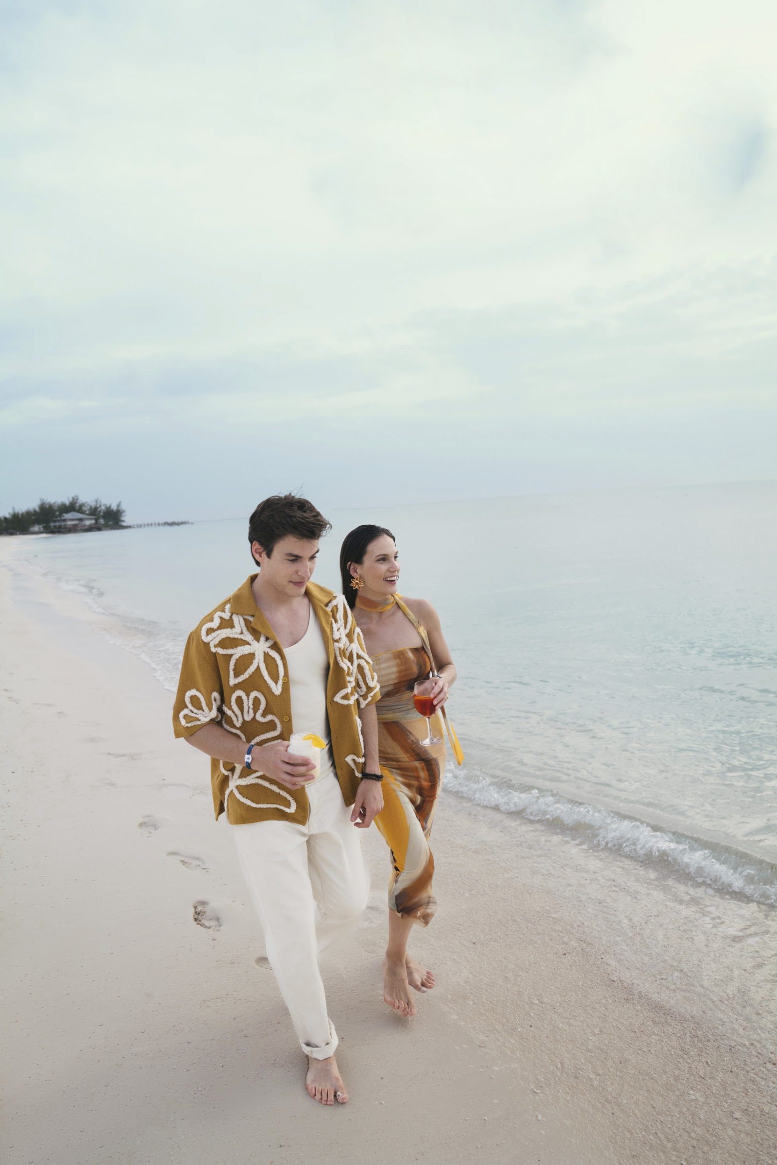 A couple walks along the beach at Club Med Columbus Isle, holding drinks with palm trees visible in the distance.