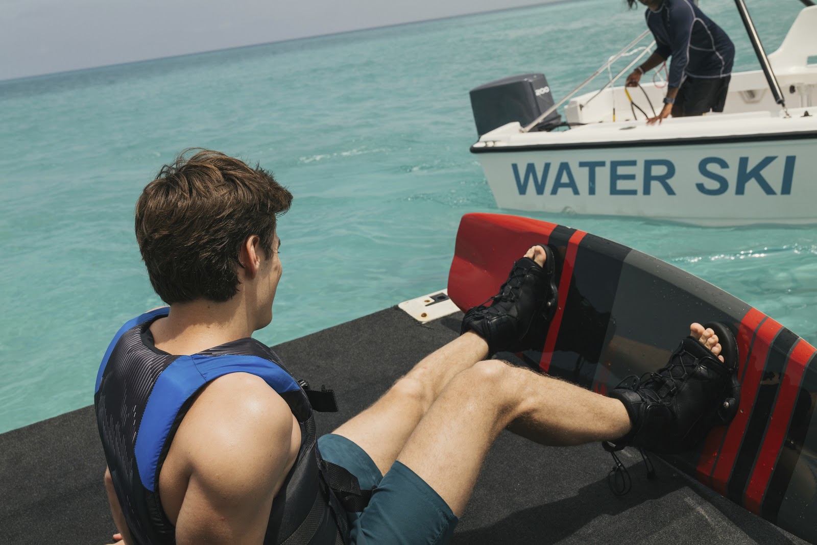 Man in life jacket sits on water ski boat with operator in turquoise Caribbean waters.