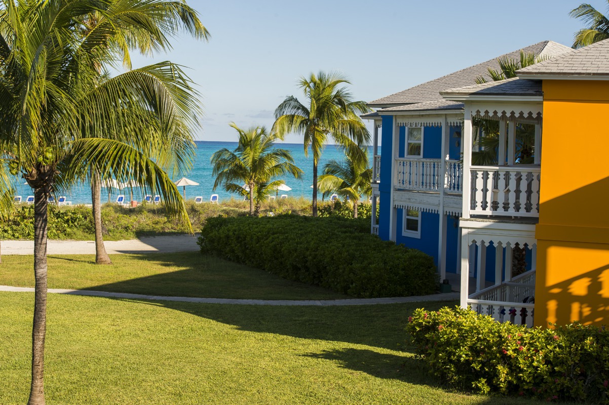 Colorful colonial-style buildings with blue and yellow facades overlook manicured lawns and palm trees toward turquoise ocean waters at Club Med Columbus Isle.