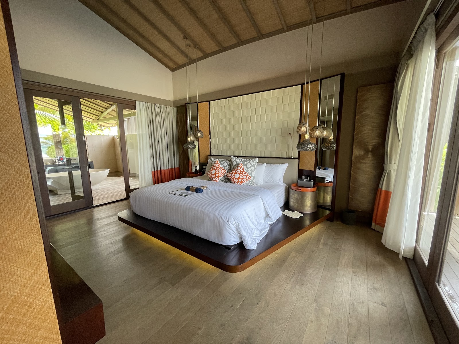 Resort bedroom with raised platform bed, white linens, orange accent pillows, pendant lights, and glass doors opening to outdoor bathtub area.