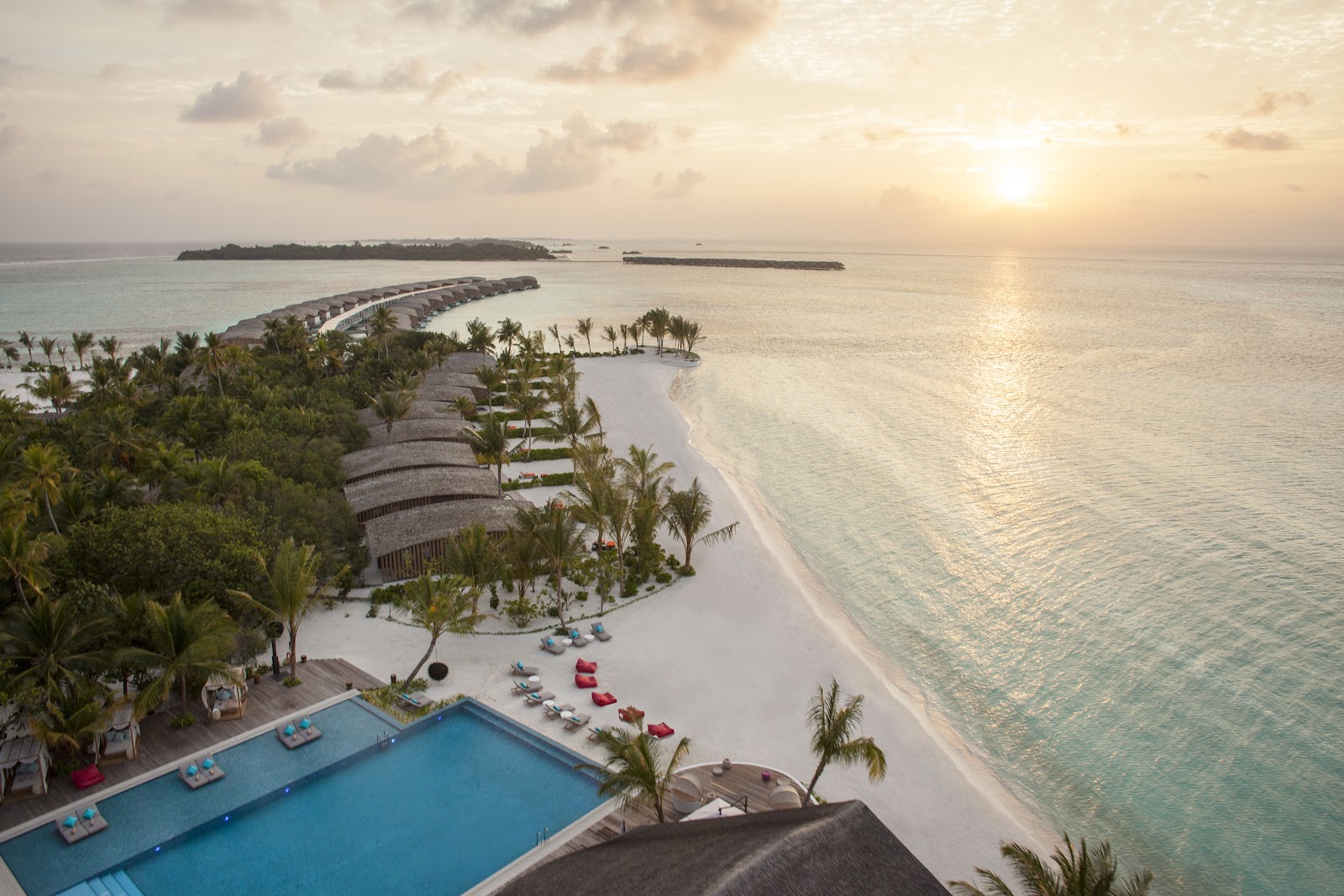 Aerial view of Club Med Finolhu Villas with curved overwater villas, palm-lined beach, blue pool, and distant islands at sunset.