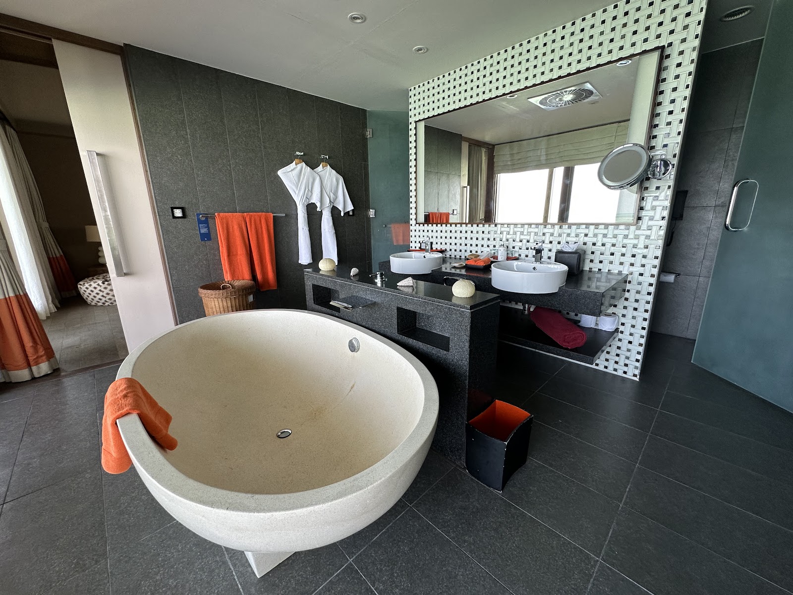 Modern resort bathroom with round soaking tub, dual sink vanity, large mirror, and dark tile floors accented with orange towels.