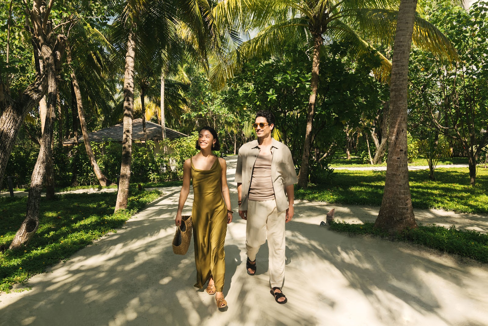 A couple walks hand in hand along a shaded path lined with tall palm trees and lush green vegetation at Club Med Finolhu Villas.