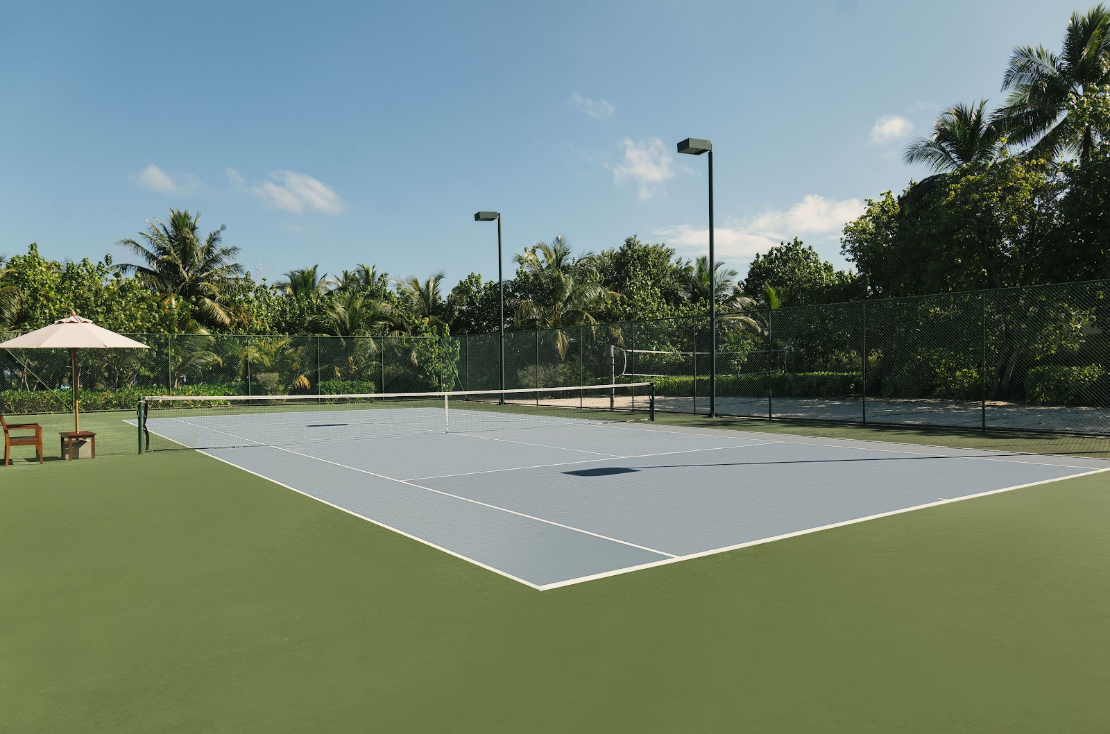Tennis courts with gray playing surfaces and white lines surrounded by palm trees and tropical vegetation under clear skies.