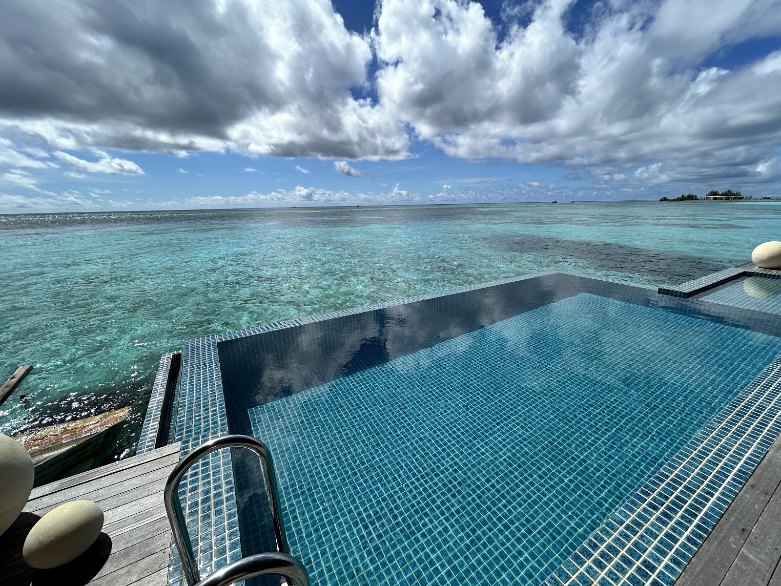 Overwater pool with blue tile deck overlooking turquoise lagoon and distant islands under cloudy sky at Club Med Finolhu Villas.