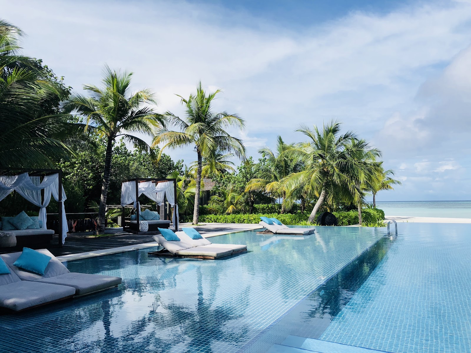 Infinity pool with lounge chairs and cabanas overlooking the ocean at Club Med Finolhu Villas, surrounded by palm trees.