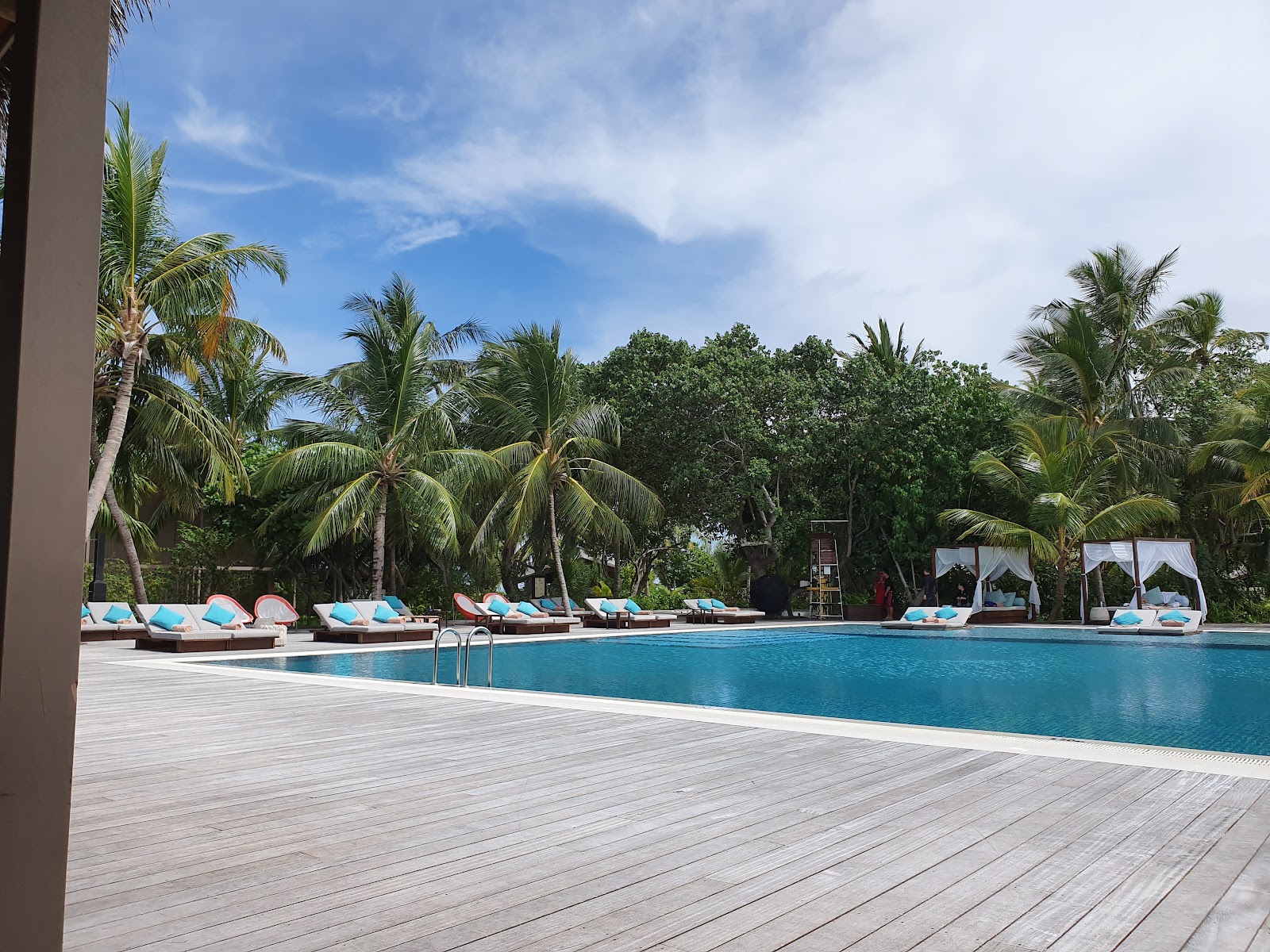 Resort pool with turquoise water, wooden deck, lounge chairs with blue cushions, palm trees, and tropical vegetation under a partly cloudy sky.