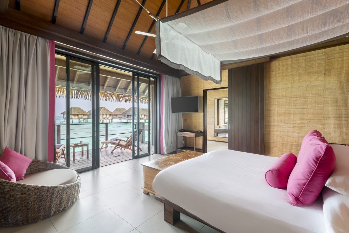 Overwater bungalow bedroom with king bed, pink pillows, and glass doors opening onto a deck with loungers and lagoon view.