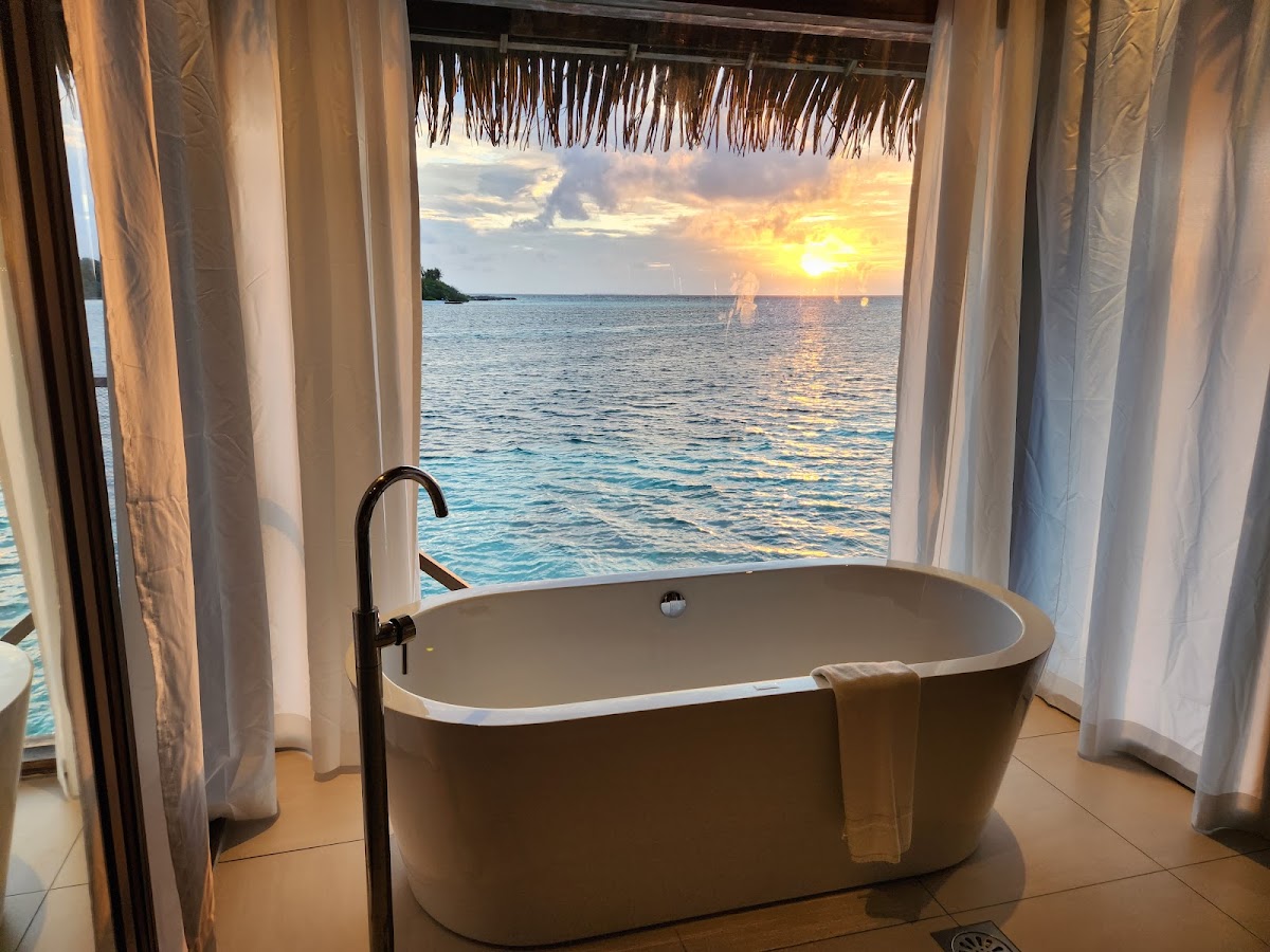 Freestanding bathtub with ocean view through open windows at sunset, thatched roof visible above.