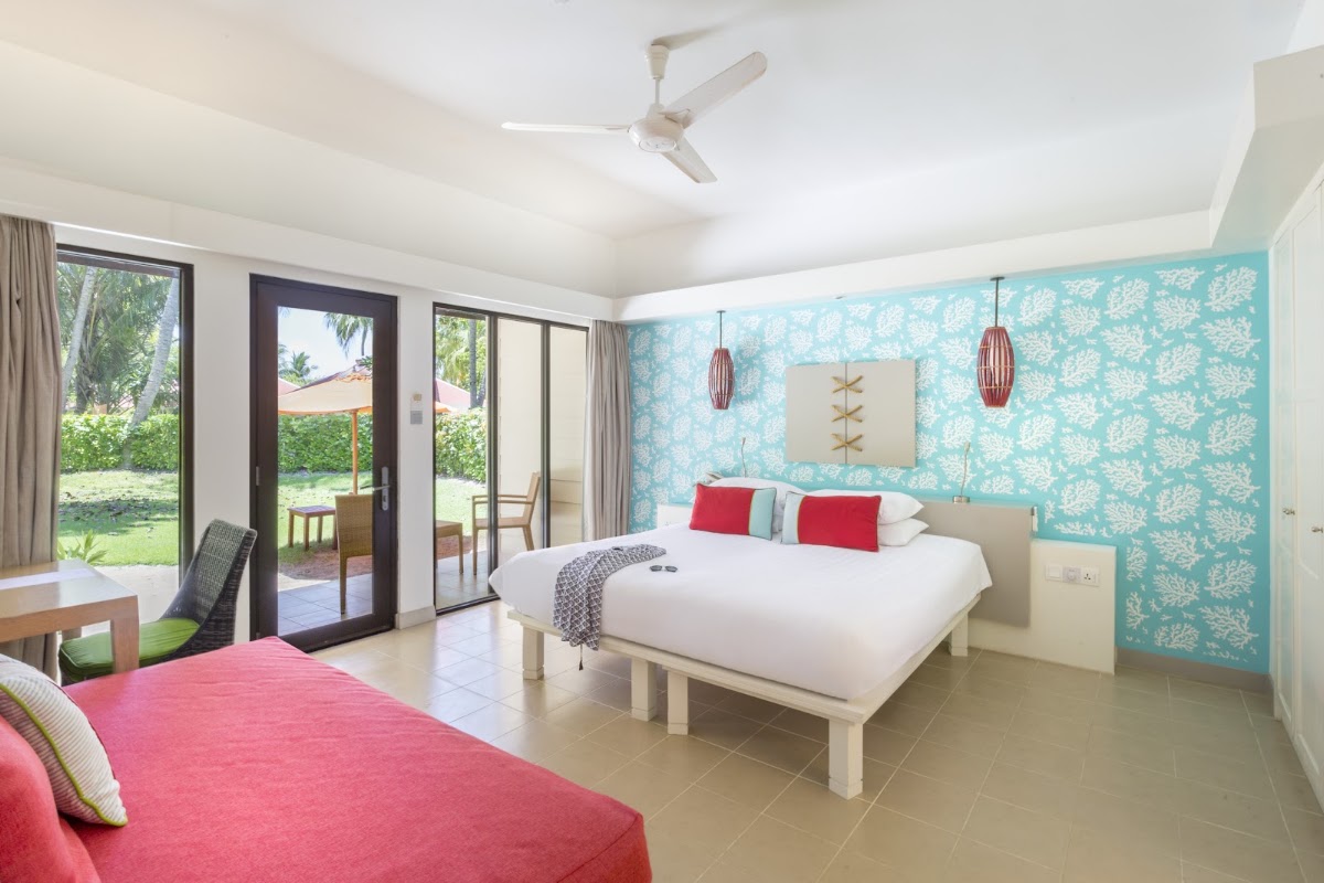 Modern resort bedroom with turquoise coral-patterned accent wall, two beds with red and white bedding, and glass doors opening to tropical gardens.