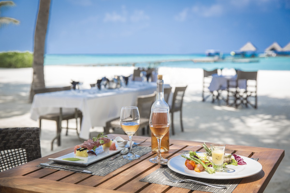 Beach dining table set with plated appetizers, wine glass and bottle, overlooking white sand and turquoise water with overwater bungalows.