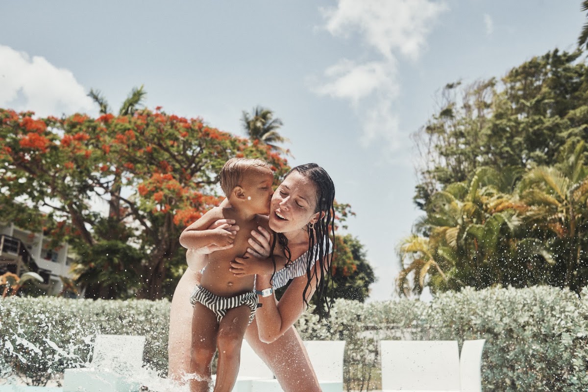 Woman and toddler splash in shallow water surrounded by tropical flowers and palm trees at a resort pool area.