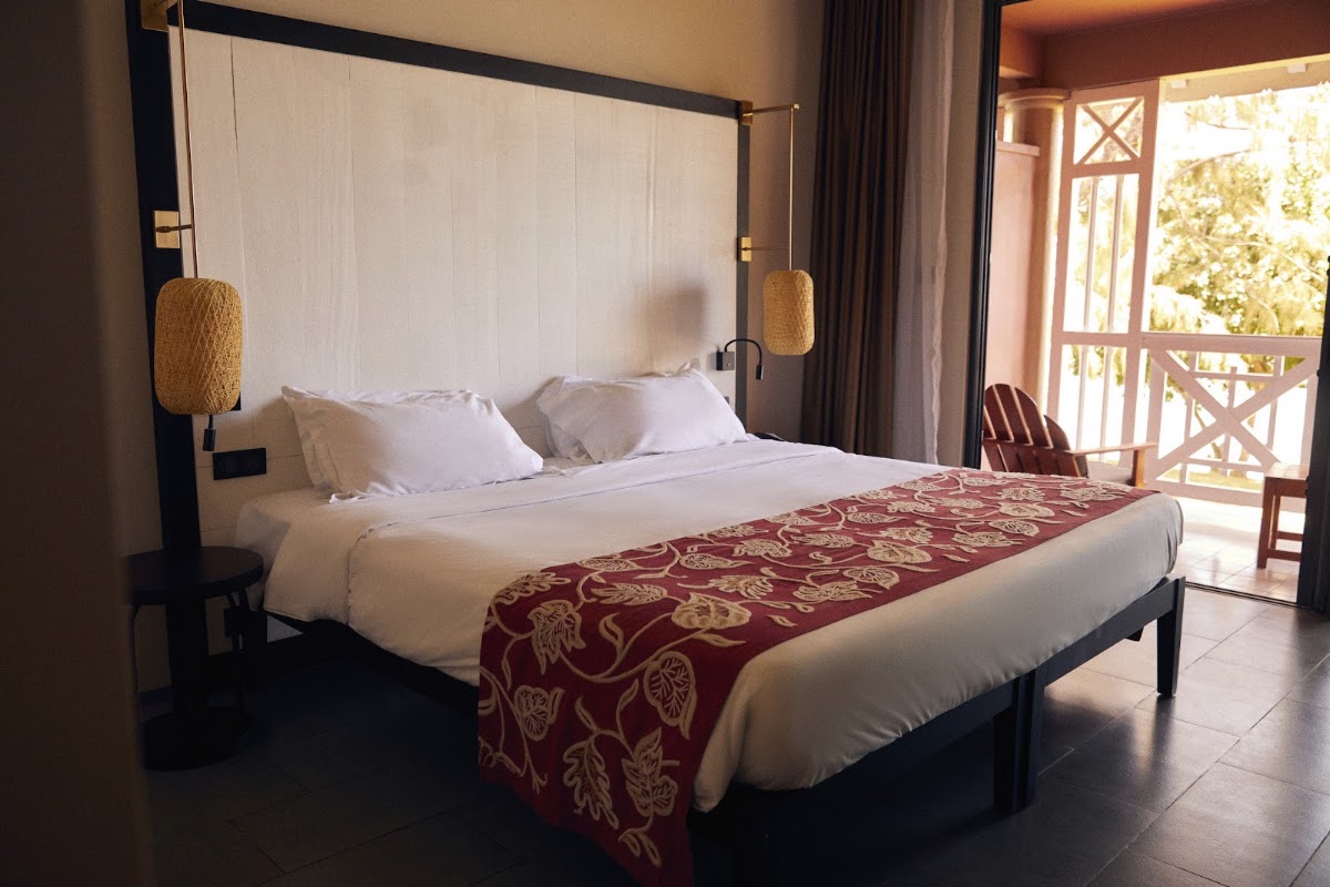 Resort room with a king bed, cream linens, burgundy patterned runner, rattan pendant lights, and doors opening to a tropical balcony.
