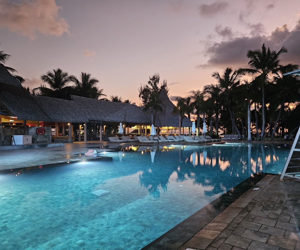 Resort pool at dusk with thatched-roof buildings, palm trees, and lounge chairs reflected in calm turquoise water.