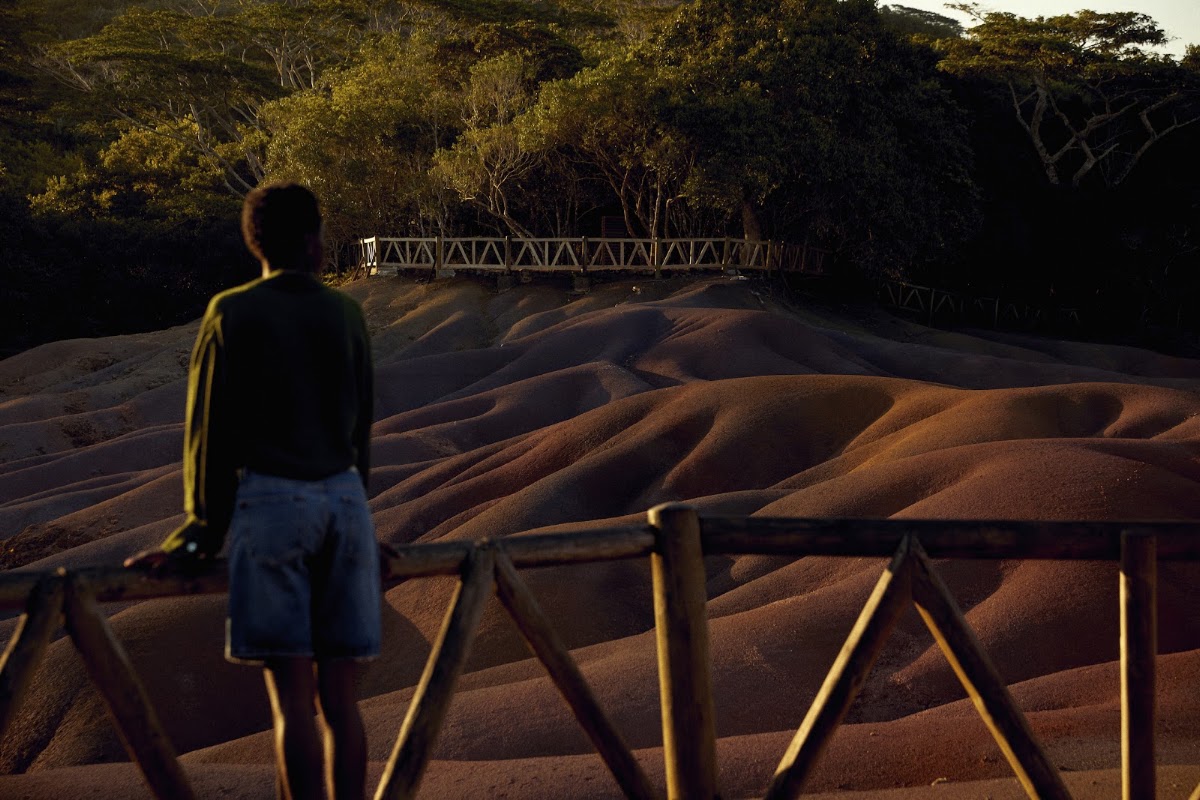 Child standing at wooden railing overlooking rippled sand dunes with dense forest and metal footbridge in background at sunset.