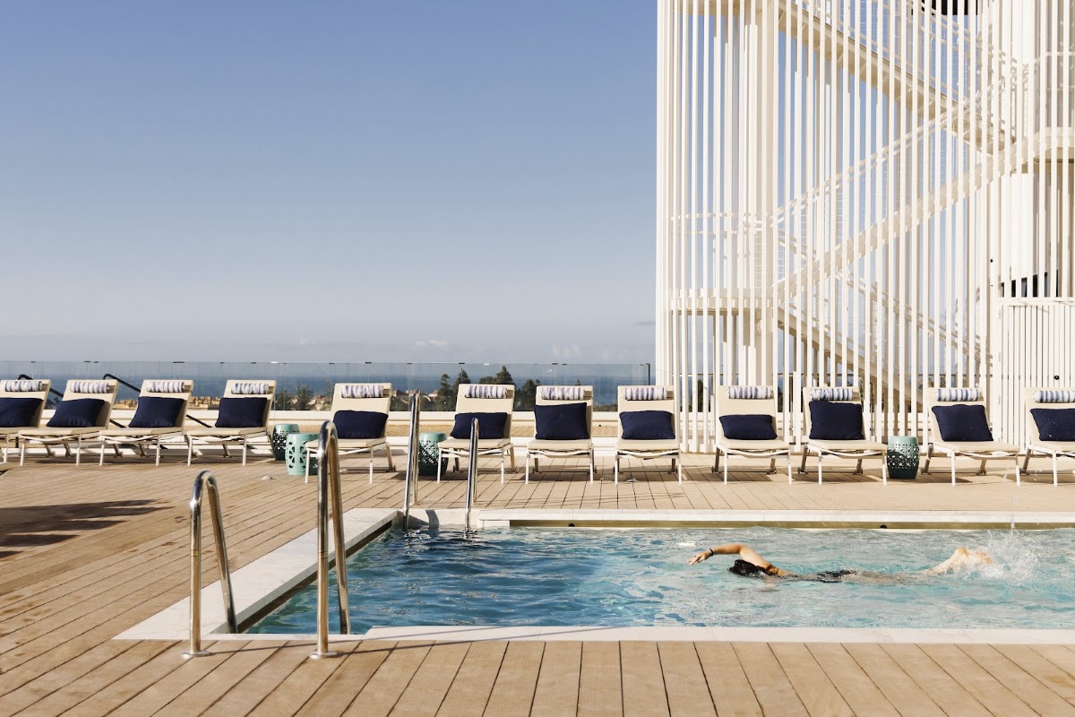 Swimmer doing laps in a wooden-deck pool with white loungers and a modern building overlooking mountains at Club Med Magna Marbella.