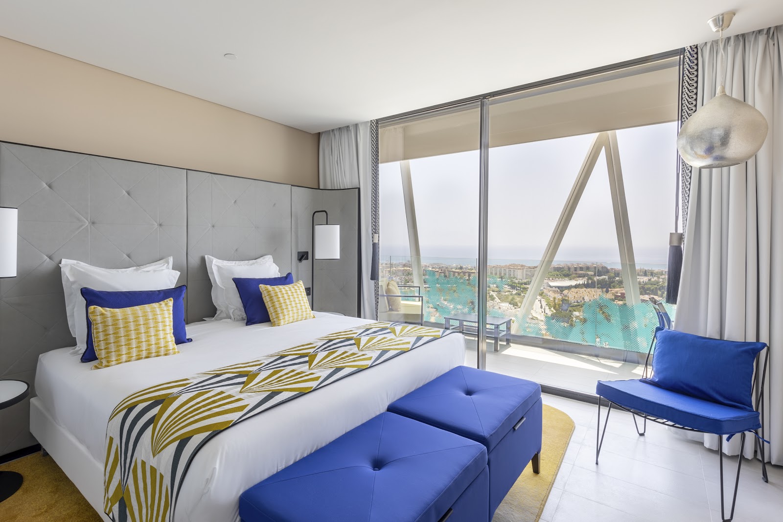 Resort bedroom with queen bed, blue and yellow accents, and floor-to-ceiling windows overlooking a turquoise lagoon and coastal landscape.