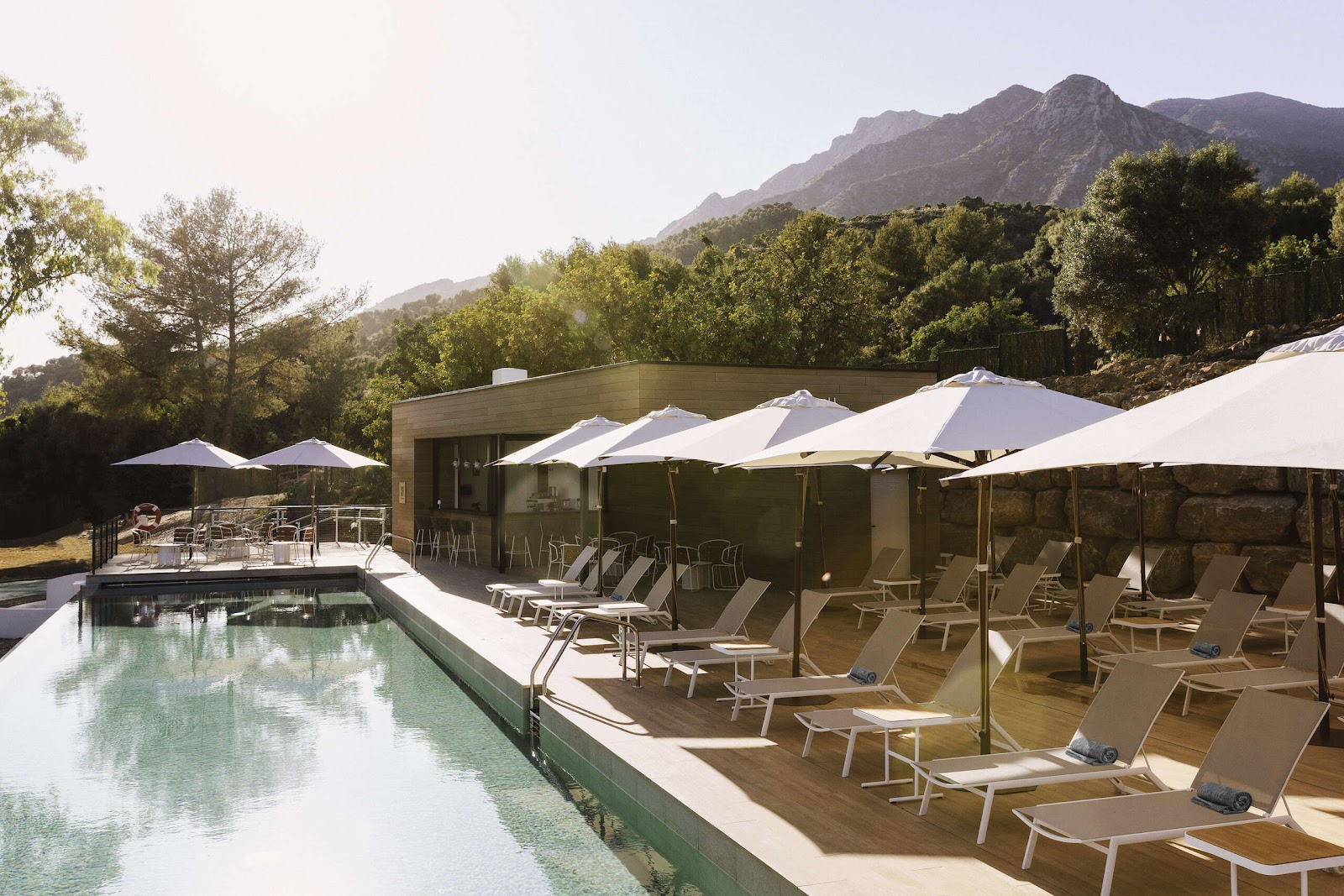 Pool deck at Club Med Magna Marbella with lounge chairs under white umbrellas, modern building, and mountains in background.