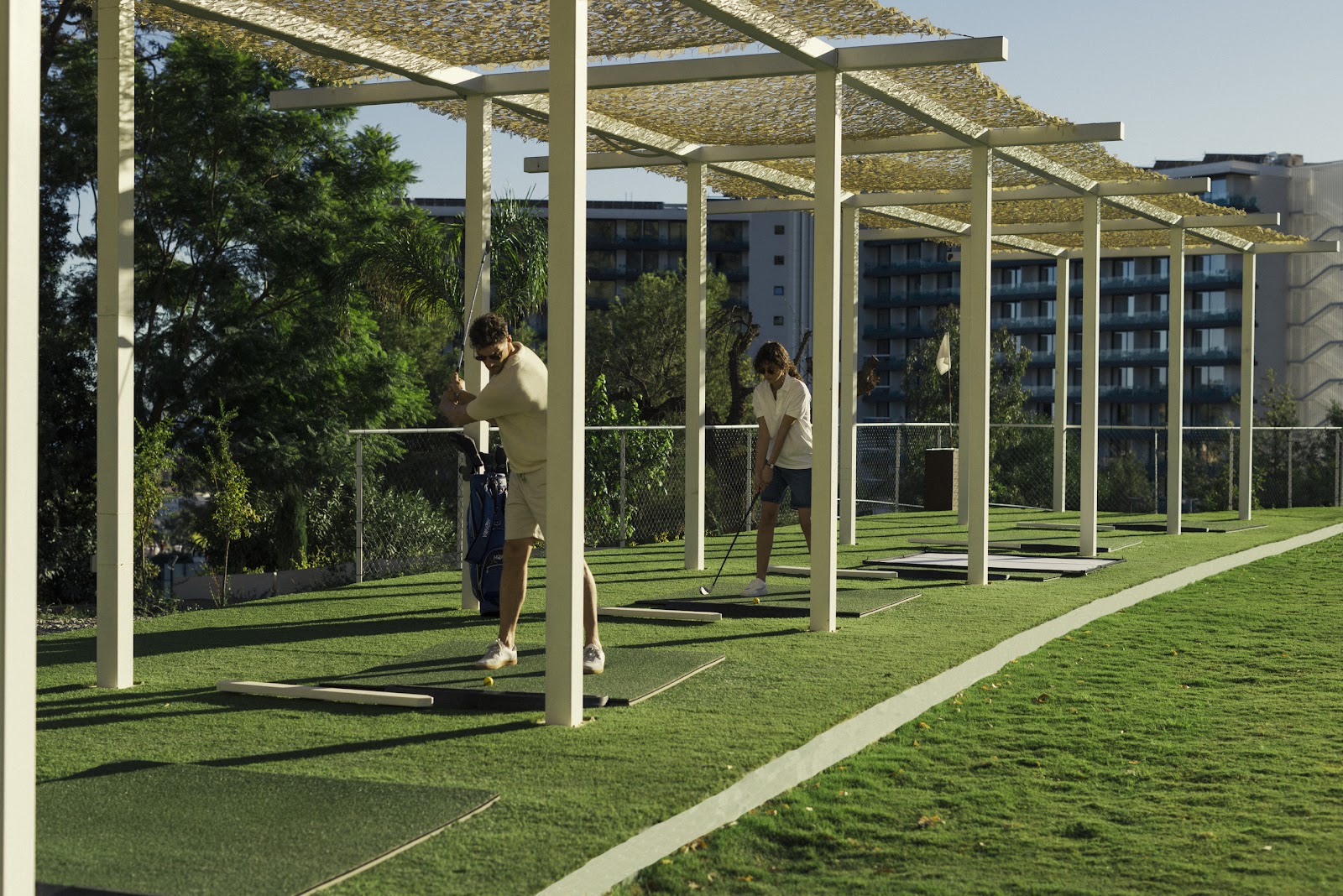 Two guests play mini golf under white pergolas with woven shade panels on a manicured lawn, with resort buildings visible beyond.
