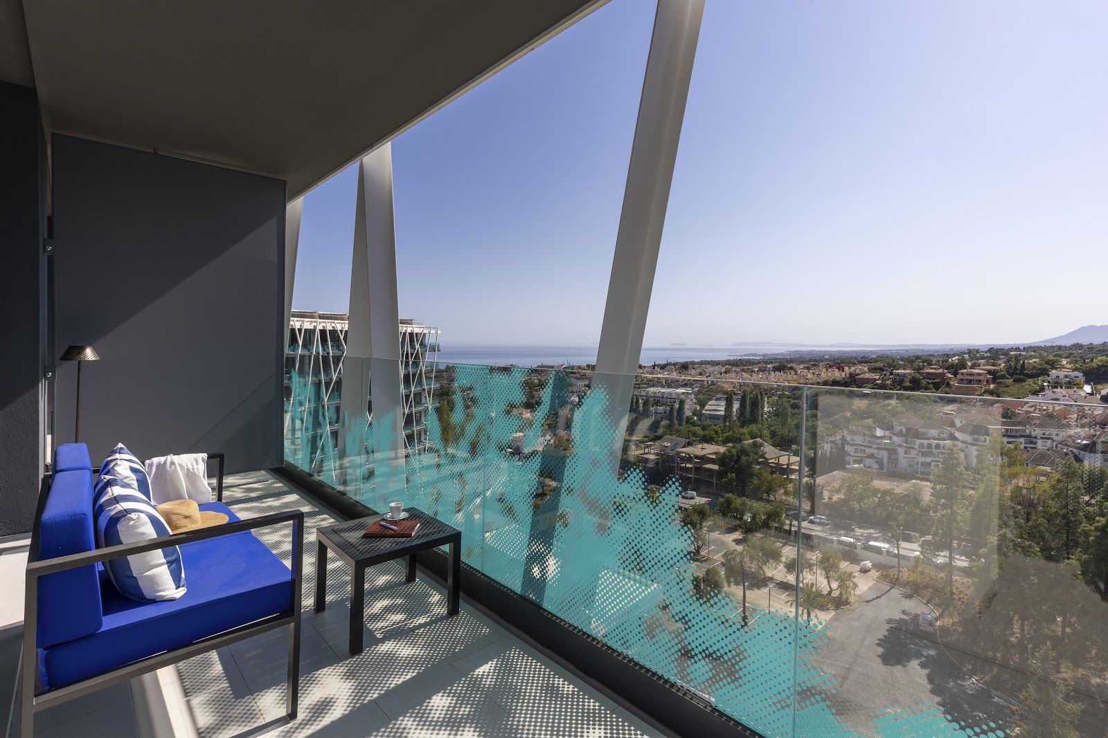 Blue lounge chair on a high-rise balcony overlooking a coastal town, sea, and mountains under clear sky.