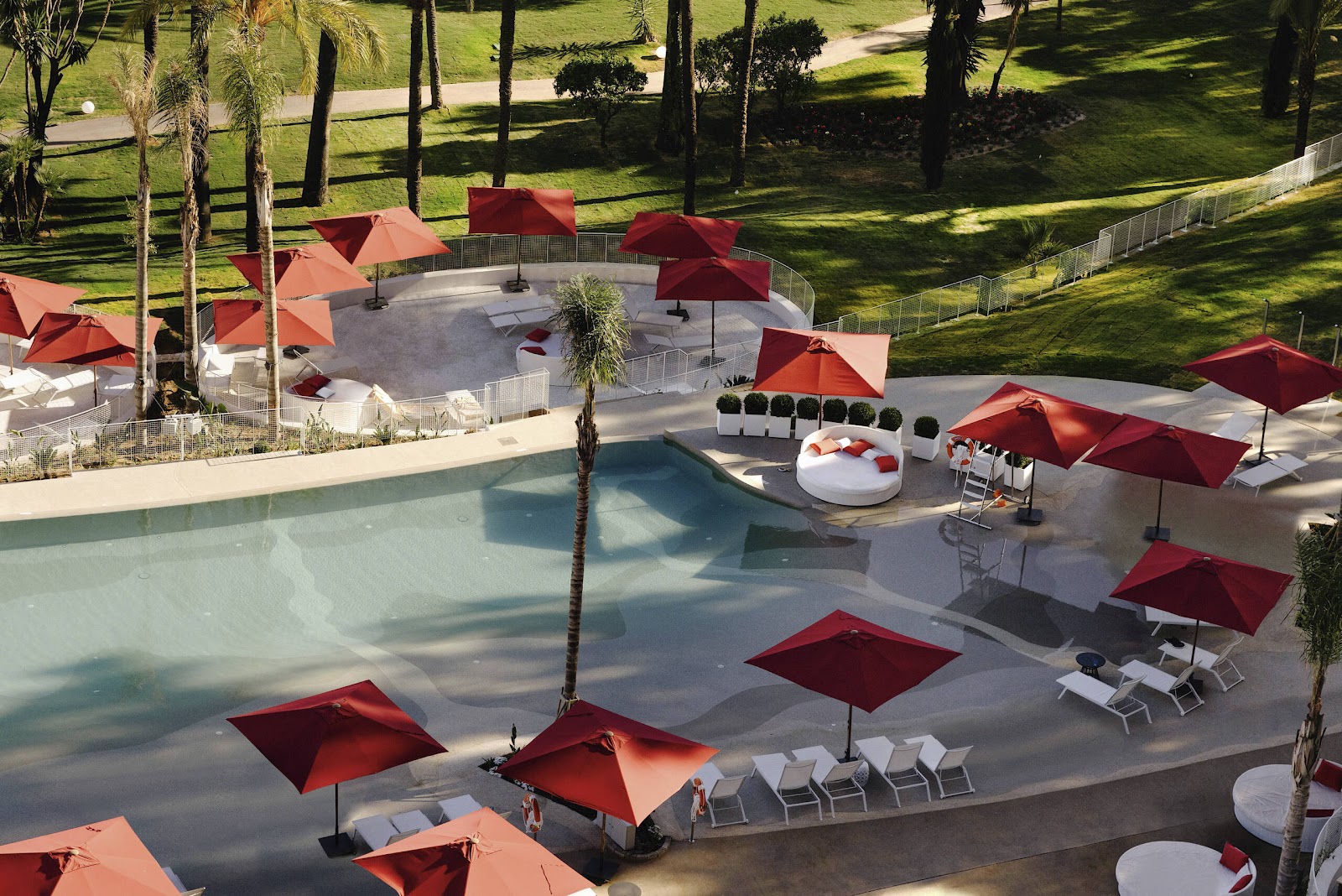 Red umbrellas and white loungers surround a pool at Club Med Magna Marbella, with manicured lawns and palm trees beyond.