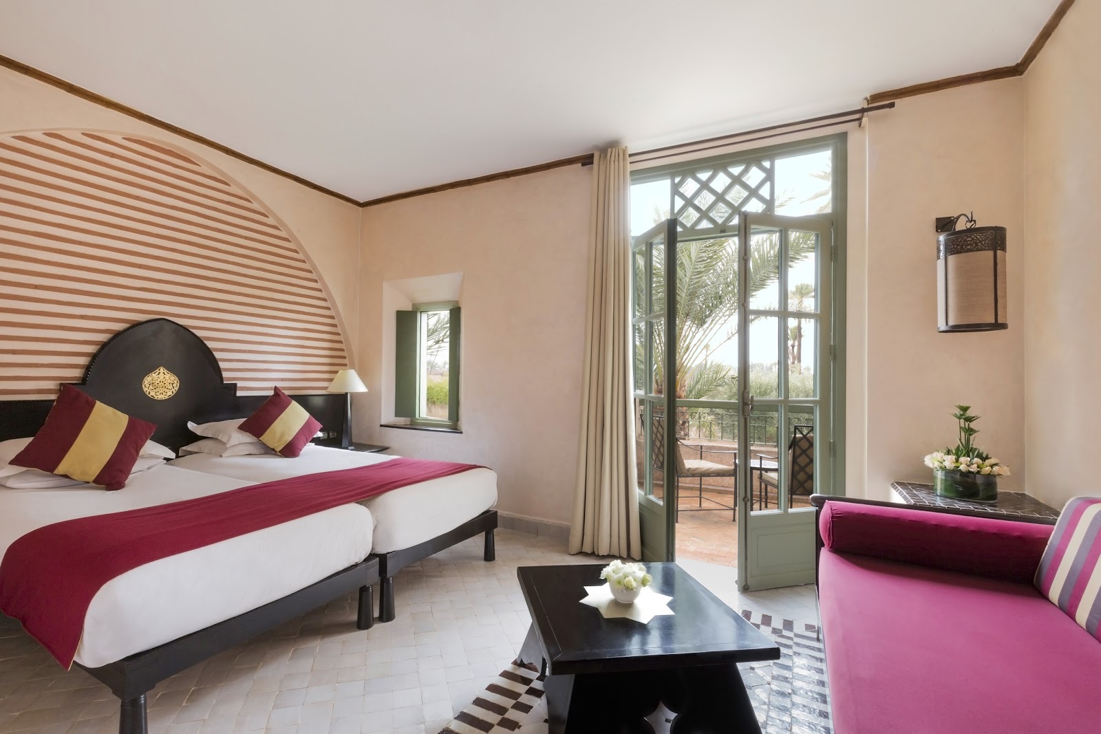 Resort bedroom with king bed, burgundy accents, pink seating area, and balcony overlooking palm trees at Club Med Marrakech La Palmeraie.